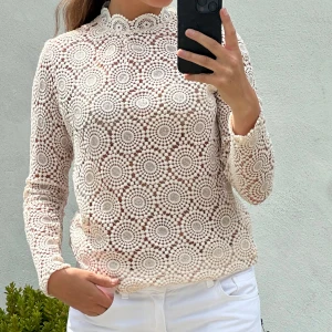 Vit spetsblus med cirkelmönster - Superfin vit spetsblus med lång ärm och högt hals med vågig kant. Blusen har ett detaljerat cirkelmönster i spets och är genombruten, vilket ger en luftig och trendig look. Perfekt att styla med jeans eller kjol för en chic vibe.