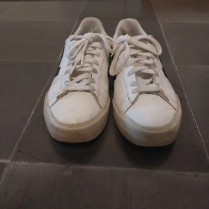 Vita Veja sneakers med svart detalj - Snygga vita sneakers från Veja med svart V-logga på sidan och svart detalj på hälen. Klassisk låg modell med rund tå, snörning och platt sula. Tillverkade i skinn för en clean och stilren look. Perfekta för dig som gillar minimalistisk stil.
