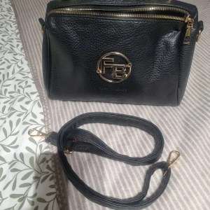 Svart axelväska från Fashion Bags - Snygg svart axelväska i skinnimitation från Fashion Bags med guldfärgade detaljer och logga framtill. Väskan har justerbar axelrem, dragkedja upptill och flera fack inuti för smidig organisering. Perfekt storlek för det viktigaste du behöver ha med dig.