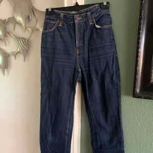 Mörkblå Nudie Jeans  - Säljer ett par mörkblå jeans från Nudie Jeans med klassiska orange sömmar och snygga bakfickor med vågig detalj. Jeansen är tillverkade i 100% ekologisk bomull och har en rak passform. Perfekta för dig som gillar stilrena och tidlösa jeans, jeansen har en liten slitning där bak som man kan se på bild två men annars är det inga konstigheter med dem 