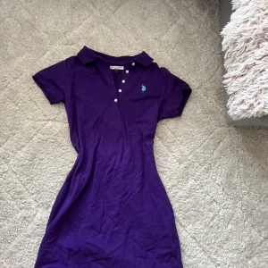 Lila kort pikéklänning från Polo Ralph Lauren - Snygg lila pikéklänning från Polo Ralph Lauren med klassisk krage, korta ärmar och knappar framtill. Klänningen har en figurnära passform och den ikoniska loggan broderad på bröstet i turkos. Perfekt för en sportig och avslappnad look.