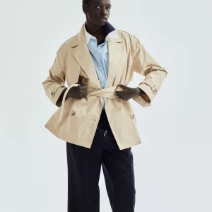 Beige trenchcoat med bälte - Snygg beige trenchcoat med bälte och knappar. Perfekt för vår och höst. Jackan har en klassisk design med krage och långa ärmar.