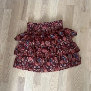 Lindex mönstrad volangkjol - Säljer en kort mönstrad volangkjol i rött och blått med elastisk midja. Kjolen har flera lager volanger och ett bohemiskt mönster som ger en unik stil. Perfekt för att lägga till lite färg och rörelse i din garderob!💞