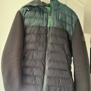 Svart och grön jacka från BOSS - Snygg jacka från BOSS i grått och grönt med dragkedja och huva. Jackan har långa ärmar och är quiltad för extra värme. Perfekt för vår dagar och sommarkvällar.