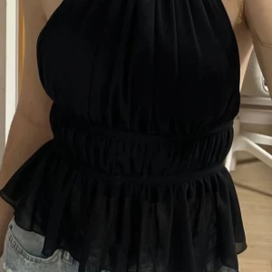 Svart halterneck topp - Snygg svart halterneck topp med öppen rygg och knytband. Toppen har en volangdetalj vid midjan och är perfekt för en somrig look. Passar bra till jeans eller kjol.