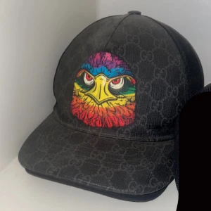 Gucci keps - Snygg svart keps från Gucci med ett coolt mönster och en färgglad örn på framsidan. Kepsen har en böjd skärm och justerbar passform. Perfekt för att ge din outfit en unik touch. Mycket svår att få tag på