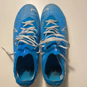 Blå Nike fotbollsskor - Säljer ett par blå Nike fotbollsskor med vit snörning och texten 'JUST DO IT' på sulan. Skorna har en strömlinjeformad design och är perfekta för snabba rörelser på planen. De har en lätt och ventilerande ovandel. Storlek 40