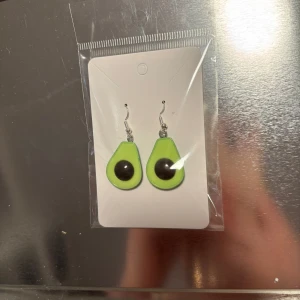 Gröna avokado-örhängen - Säljer ett par roliga örhängen i form av avokado. De är gröna med en brun kärna och har en krok för enkel användning. Perfekta för att ge en färgglad touch till din outfit! Finns 2 par. (Billigt material) 