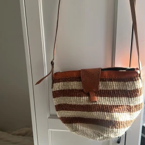 Randig axelväska i naturmaterial - Snygg axelväska med randigt mönster i brunt och beige. Väskan har en justerbar axelrem och stängs med en läderflik. Perfekt för en avslappnad stil.