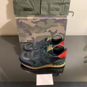 Valentino Rockrunner sneakers i camouflage - Snygga Valentino Rockrunner sneakers i en cool mix av grönt och blått camouflage. Skorna har röda detaljer på hälen och en unik design med snörning. Perfekta för dig som vill sticka ut med stil. Dustbag, kort, påse till kommer 