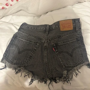 Svarta jeansshorts från Levi's - Säljer ett par coola svarta jeansshorts från Levi's med fransiga kanter och slitna detaljer. De har en knappgylf och klassiska femficksdesign. Perfekta för en avslappnad stil!