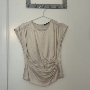 Beige satinblus med drapering - Elegant beige blus i satin med draperad detalj på sidan. Blusen har en lös passform och korta ärmar, perfekt för en stilren look. Den mjuka glansen ger en lyxig känsla.