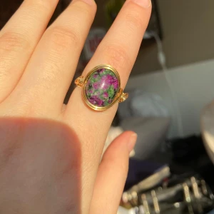 Guldring med lila och grön sten - Elegant guldring med en stor oval sten i lila och gröna nyanser. Stenen är infattad i en enkel men stilfull guldram som ger ringen en lyxig känsla. Perfekt för att ge en färgklick till din outfit. - Nickelfri - Non-tarnish behandling - Storleksguide finns på sista bilden 