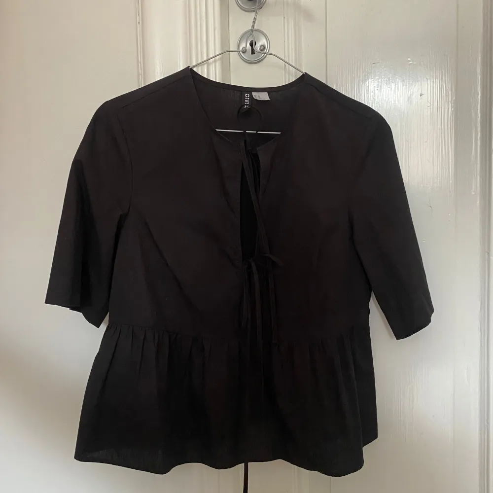 Säljer en stilren svart blus med knytning framtill. Blusen har trekvartsärmar och en lätt volangdetalj vid nederkanten. Perfekt för en chic och avslappnad look.. Yläosat.