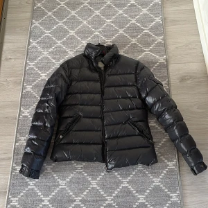 Svart dunjacka från Moncler - Säljer en snygg svart dunjacka från Moncler. Jackan har en glansig finish med dragkedja och hög krage. Den har långa ärmar och praktiska fickor med dragkedja. Perfekt för kyliga dagar!