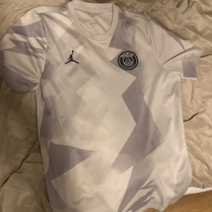 Vit och grå PSG fotbollströja - Snygg PSG fotbollströja i vitt och grått med ett coolt geometriskt mönster. Tröjan har korta ärmar och PSG-loggan på bröstet tillsammans med Jordan-loggan. Perfekt för fotbollsfans som vill ha en stilren look.