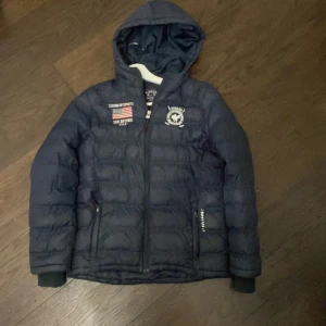 Mörkblå pufferjacka från Vinson Polo Club - Säljer en mörkblå pufferjacka från Vinson Polo Club med huva och dragkedja. Jackan har broderade detaljer på bröstet och ribbade muddar vid ärmsluten. Perfekt för kyliga dagar!