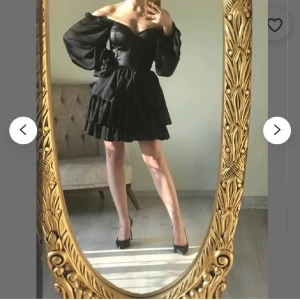 Svart off-shoulder klänning med volanger - Snygg svart off-shoulder klänning med volanger och snörning i ryggen. Klänningen har långa puffärmar och en kort, flerdelad kjol som ger en lekfull look. Perfekt för en kväll ute eller en speciell tillställning.