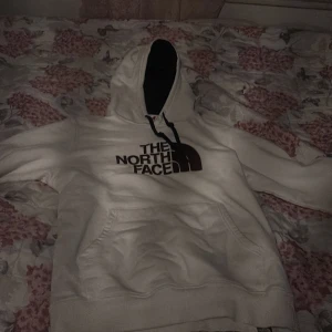 Vit hoodie från The North Face - Säljer en vit hoodie från The North Face med deras ikoniska logga i svart och brunt på framsidan. Hoodien har en klassisk känguruficka och en justerbar huva med snören. Perfekt för en avslappnad stil.