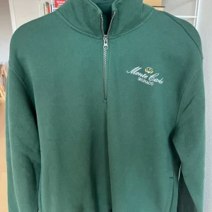 Grön tröja med dragkedja från Hollister - Säljer en grön tröja från Hollister med halv dragkedja och texten 'Monte Carlo Monaco' broderad på bröstet. Perfekt för en avslappnad stil.