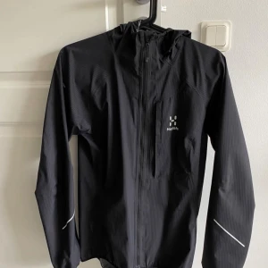  Haglöfs vindjacka & regnjacka -kvinna - Säljer en stilren svart vindjacka från Haglöfs med dragkedja och huva. Jackan har långa ärmar och reflekterande detaljer för extra synlighet. Perfekt för utomhusaktiviteter i blåsigt väder. Storlek S men funkar även som M. (Kvinnlig modell). 