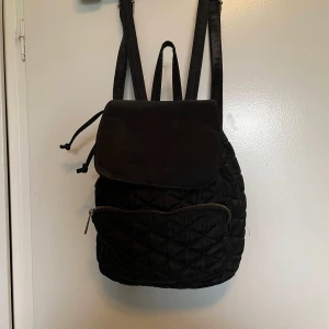 Svart quiltad ryggsäck - Snygg svart quiltad ryggsäck med dragkedja och snörning. Perfekt för att bära dina saker med stil. Den har en praktisk ficka framtill och justerbara axelremmar för bekväm användning.