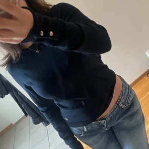 Mörkblå tröja från Holly & Whyte - Säljer en tajt mörkblå tröja från Holly & Whyte by Lindex. Tröjan har långa ärmar med knappar vid ärmsluten och en ribbad kant vid halsen. Perfekt för en elegant och avslappnad look.