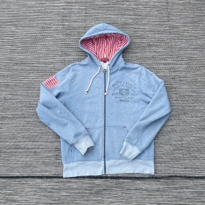 Blå hoodie från Polo Jeans Company - Säljer en snygg blå hoodie från Polo Jeans Company med dragkedja och randigt foder i huvan. Den har en amerikansk flagga på ärmen och tryck på bröstet. Perfekt för sommaren🔥🔥Priset kan diskuteras 🙌