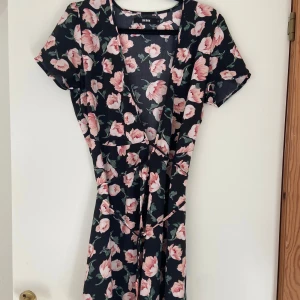 Blommig klänning från Bik Bok - Söt blommig klänning från Bik Bok med rosa blommor på en mörk bakgrund. Klänningen har korta ärmar och ett knytband i midjan för en fin passform. 