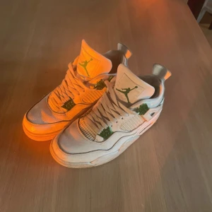 Nike Air Jordans 4 med gröna detaljer - Snygga Nike Air Jordans i vitt med gröna detaljer. Skorna har en klassisk design med snörning och den ikoniska Jumpman-loggan. Perfekta för dig som vill ha en stilren look med en touch av färg.