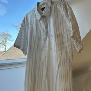 Vit och blå randig skjorta från L.G. Classic - Säljer en snygg vit skjorta med blå ränder från L.G. Klassisk, tillverkad i Italien. Skjortan har korta ärmar och en klassisk krage med knappar framtill. Perfekt för en sommarlook.