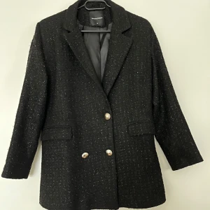 Tiffany & Tomato svart kavaj, storlek M - Glittrig svart kavaj med dubbelknäppning och guldiga knappar från Tiffany & Tomato, storlek M. Kavajen är ny oanvänd. Dock saknas ena knappen på höger sida (se bild). 100% polyester.   Mått: Längd ca 80 cm Byst ca 46 cm Midja ca 43 cm Ärmlängd mätt från axeln ca 55 cm Axelmått ca 41 cm