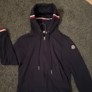 Mörkblå hoodie från Moncler - Pris kan diskuteras‼️                                          Snygg mörkblå hoodie från Moncler med dragkedja och huva. Den har vita och röda ränder på ärmslut och huva, samt Moncler-logga på ärmen. Perfekt för en stilren och bekväm look. Storlek S, men passar även XS‼️