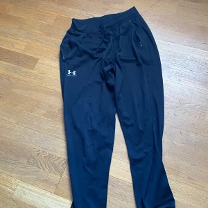 Svarta mjukisbyxor från Under Armour - Säljer ett par svarta mjukisbyxor från Under Armour med dragsko i midjan och logotyp på benet. Byxorna har en avslappnad passform och är perfekta för träning eller en chill dag hemma.