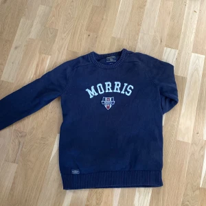 Mörkblå tröja från Morris Gents - Säljer en snygg mörkblå tröja från Morris Gents med vit text och ett emblem på bröstet. Tröjan har långa ärmar och ribbade muddar vid ärmslut och nederkant. Perfekt för en stilren look!