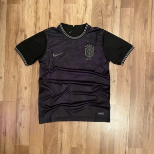 Svart Brasilien fotbollströja från Nike - Säljer en stilren svart Brasilien fotbollströja från Nike med korta ärmar. Tröjan har ett diskret mönster och det brasilianska fotbollsförbundets emblem på bröstet. Perfekt för fotbollsfans som vill ha en unik look.