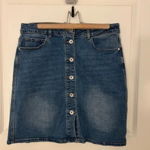 Jeanskjol med knappar - Snygg jeanskjol i klassisk blå denim med knappar framtill. Kjolen har en normal passform och praktiska fickor både fram och bak. Stretch i tyget! Aldrig använd.
