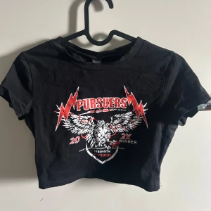 Svart croppad t-shirt från Shein - Säljer en svart croppad t-shirt från Shein med ett coolt tryck av en örn och texten 'Pursuers' i rött och vitt. Perfekt för en edgy look. T-shirten har korta ärmar och rund halsringning.