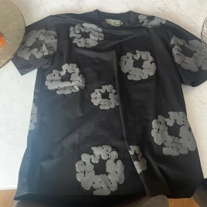 Svart t-shirt med blommönster från Denim Tears - Snygg svart t-shirt från Denim Tears med ett unikt grått blommönster. Perfekt för dig som vill ha något stilrent men ändå med en twist. Kortärmad och i mjukt material för en bekväm känsla.