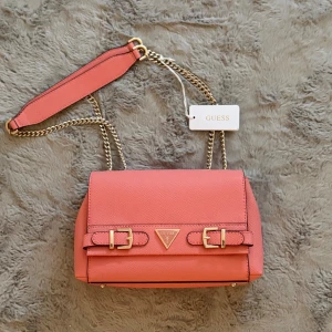 Rosa axelväska från Guess - Snygg rosa axelväska från Guess med gulddetaljer och kedjeaxelrem. Väskan har ett stilrent design med två dekorativa spännen framtill och Guess-logotypen i mitten. Perfekt för att ge en färgklick till din outfit!
