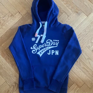 Blå hoodie från SuperDry - Säljer en snygg blå hoodie från SuperDry med vit text på framsidan. Den har en klassisk känguruficka och justerbar huva med vita snören. Perfekt för en avslappnad stil. Storlek M men passar S