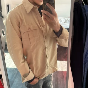 Beige overshirt från Whyred - Säljer en helt ny och stilren beige overshirt från Whyred. Skjortan har långa ärmar och knappar framtill samt två bröstfickor med lock. Perfekt för en avslappnad men ändå snygg look. Köpt för 1500kr men säljes bara för 999kr!!! Vid frågor och funderingar så är det bara att hojta till ;)