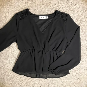 Svart blus från Vila - Snygg svart blus från Vila med v-ringning och spetsdetaljer på axlarna. Blusen har långa ärmar och en omlott-design som ger en elegant look. Perfekt för både vardag och festliga tillfällen.