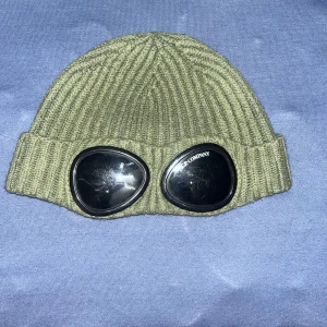 Grön ribbad mössa med glasögondetalj - Snygg grön ribbad mössa med inbyggda glasögon på framsidan. Perfekt för att sticka ut i mängden och hålla sig varm samtidigt. Glasögonen ger en unik och cool look.