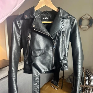 Svart skinnjacka från Zara - Snygg svart skinnjacka från Zara med dragkedja och bälte. Perfekt för en cool look.