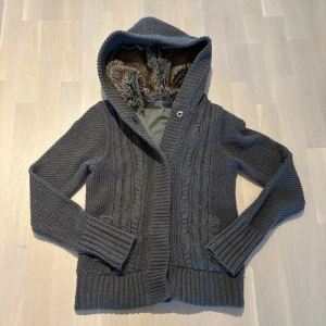 Cardigan - Säljer en mysig grå stickad kofta med pälsfoder i luvan. Tröjan har ett flätmönster framtill och ribbade muddar vid ärmslut och nederkant. Perfekt för kyliga dagar!