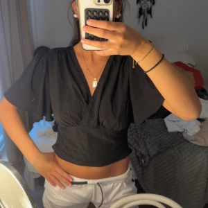 Svart omlottopp med vida ärmar - Säljer en svart omlottopp med djup v-ringning och bred resår i midjan. Toppen har korta, vida ärmar som ger en luftig känsla och croppad passform. Perfekt för dig som gillar stilrena och trendiga plagg med snygg siluett. Storlek L men känns som en M/S