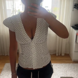 Vit prickig blus med v-ringning - Supersöt vit blus med svarta prickar och djup v-ringning. Puffiga korta ärmar och figurnära passform ger en trendig siluett. Perfekt för dig som gillar en feminin och lekfull stil. Knappar framtill och lätt stretchigt material.