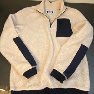 Beige fleece-tröja Jack & Jones - Mysig beige fleece-tröja från Jack & Jones med marinblå detaljer på ärmar, bröstficka och nederkant. Tröjan har half zip och hög krage, perfekt för kyliga dagar. Långärmad och med en praktisk ficka med dragkedja på bröstet.