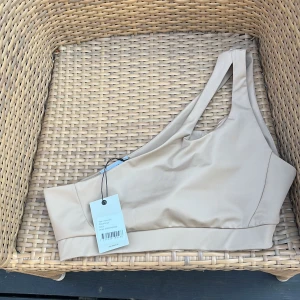 Beige one shoulder topp - Snygg beige topp med one shoulder-design och bred resår nedtill. Toppen är ärmlös och har en cool asymmetrisk skärning som ger en modern vibe. Perfekt att styla med höga jeans eller kjol för en trendig look.